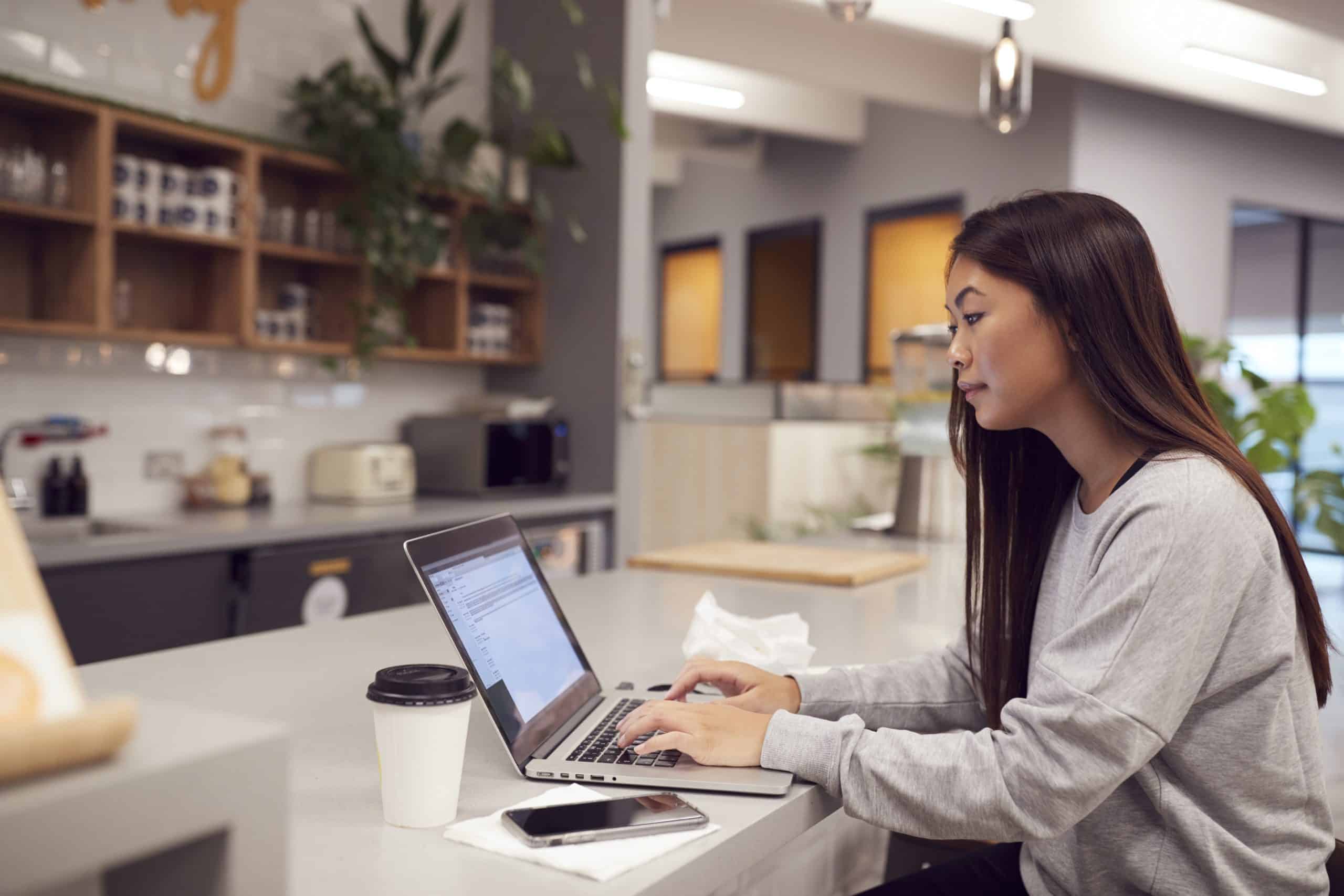 Businesswoman Working On Laptop In Kitchen Area Of Modern Office