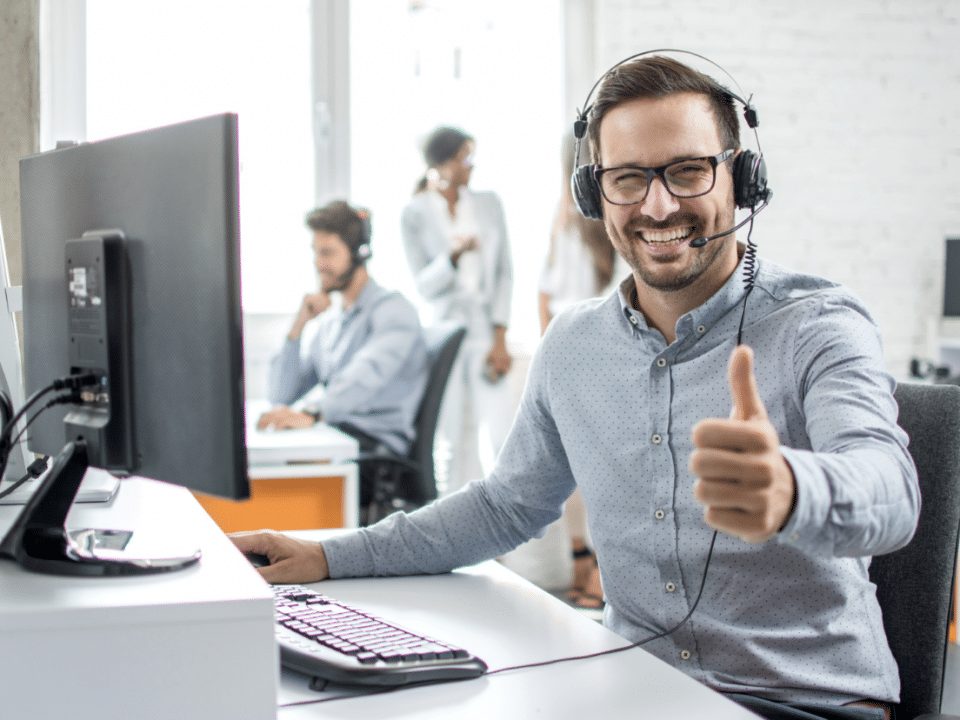 Male virtual assistant with headphones in front of computer giving thumbs up