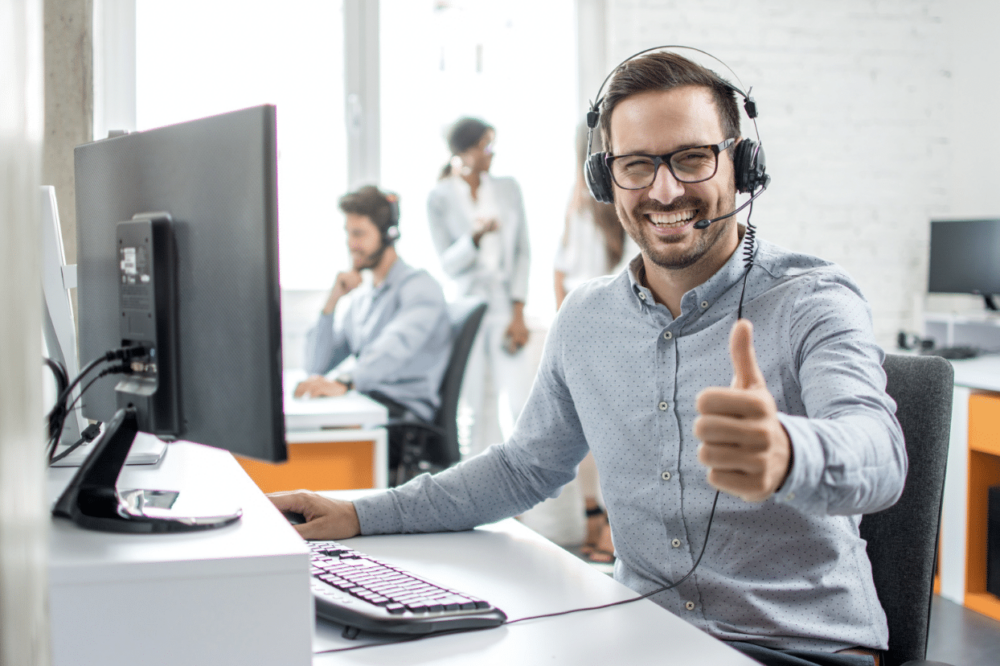 Male virtual assistant with headphones in front of computer giving thumbs up
