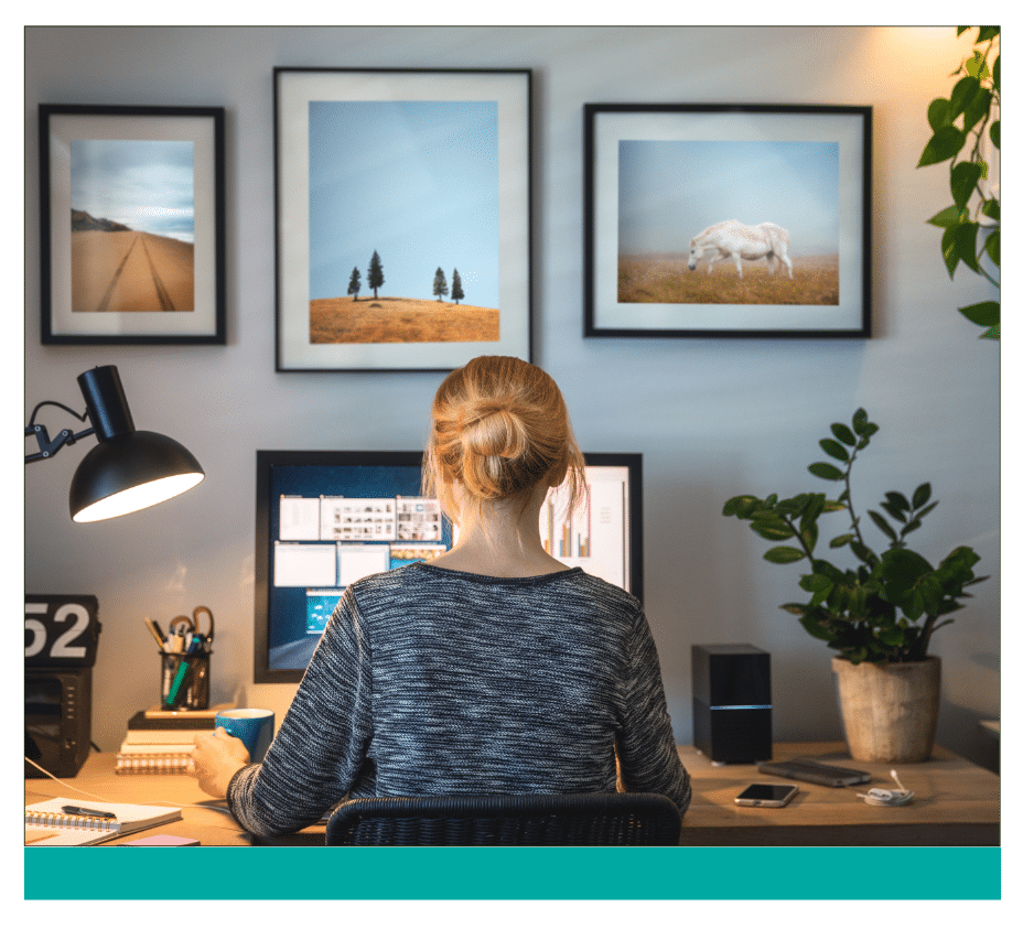89 ‣ NuvoDesk Coworking Woman working from home at a neatly organized desk with dual monitors, framed landscape art on the wall, a desk lamp, plants, and office supplies creating a cozy and productive workspace.