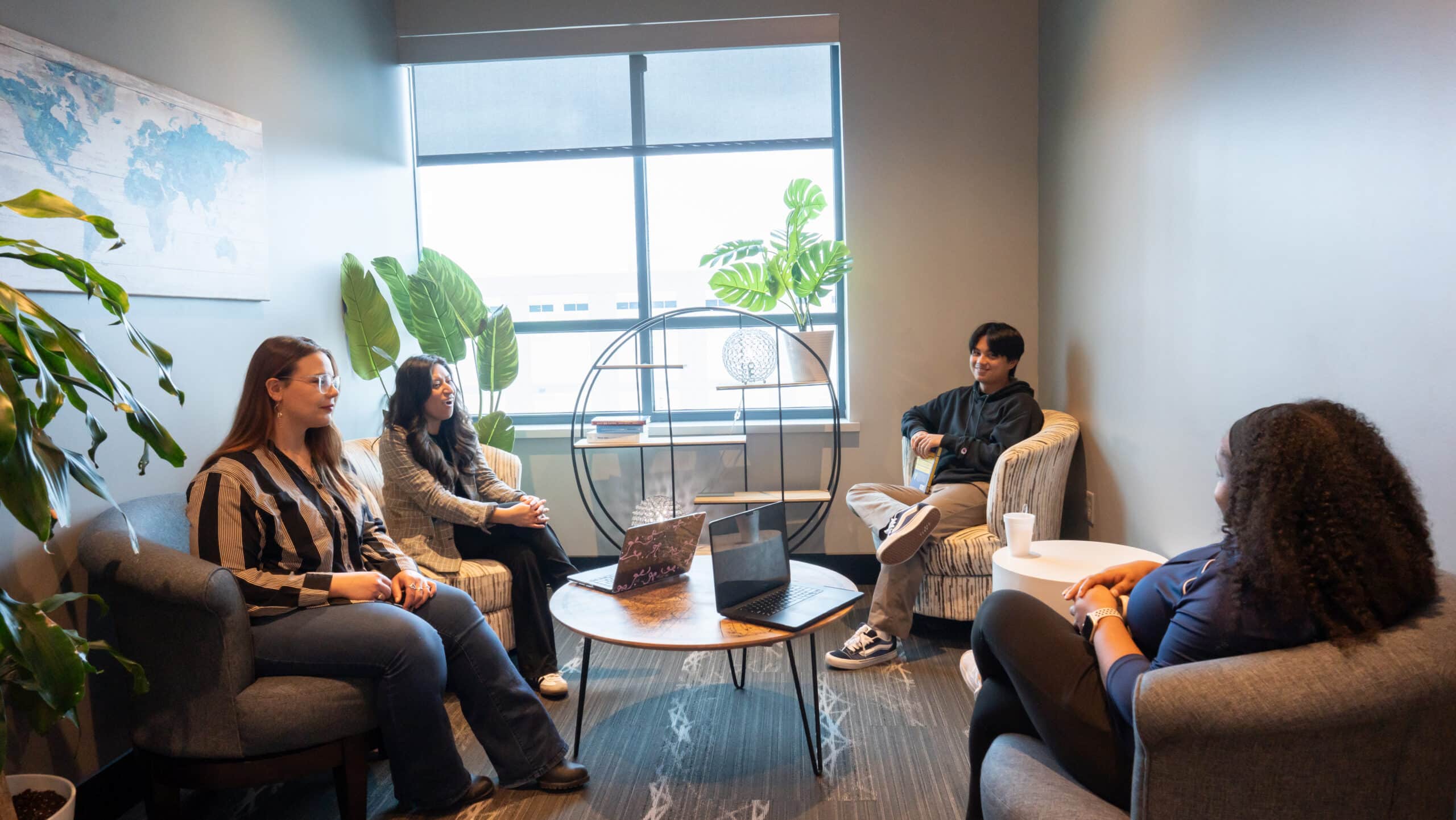 Group of five people seated in a modern lounge area with laptops, chatting by a coffee table and leafy plants near a window.