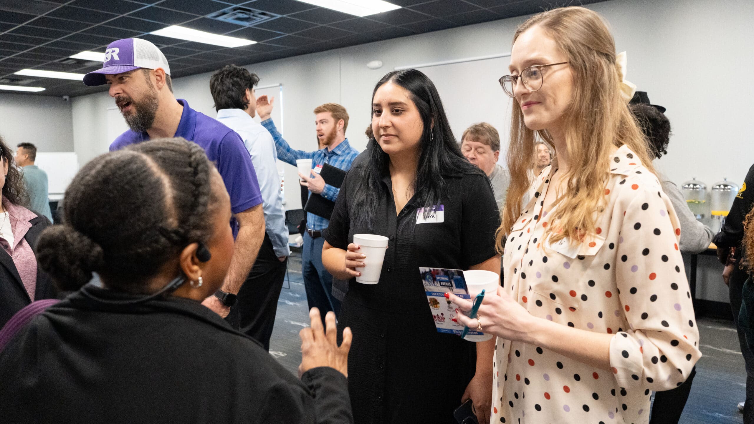 Group of people at a casual networking event, two women in foreground talking; one in a black dress holding a white cup, the other in a polka-dot blouse holding a flyer and phone case.
