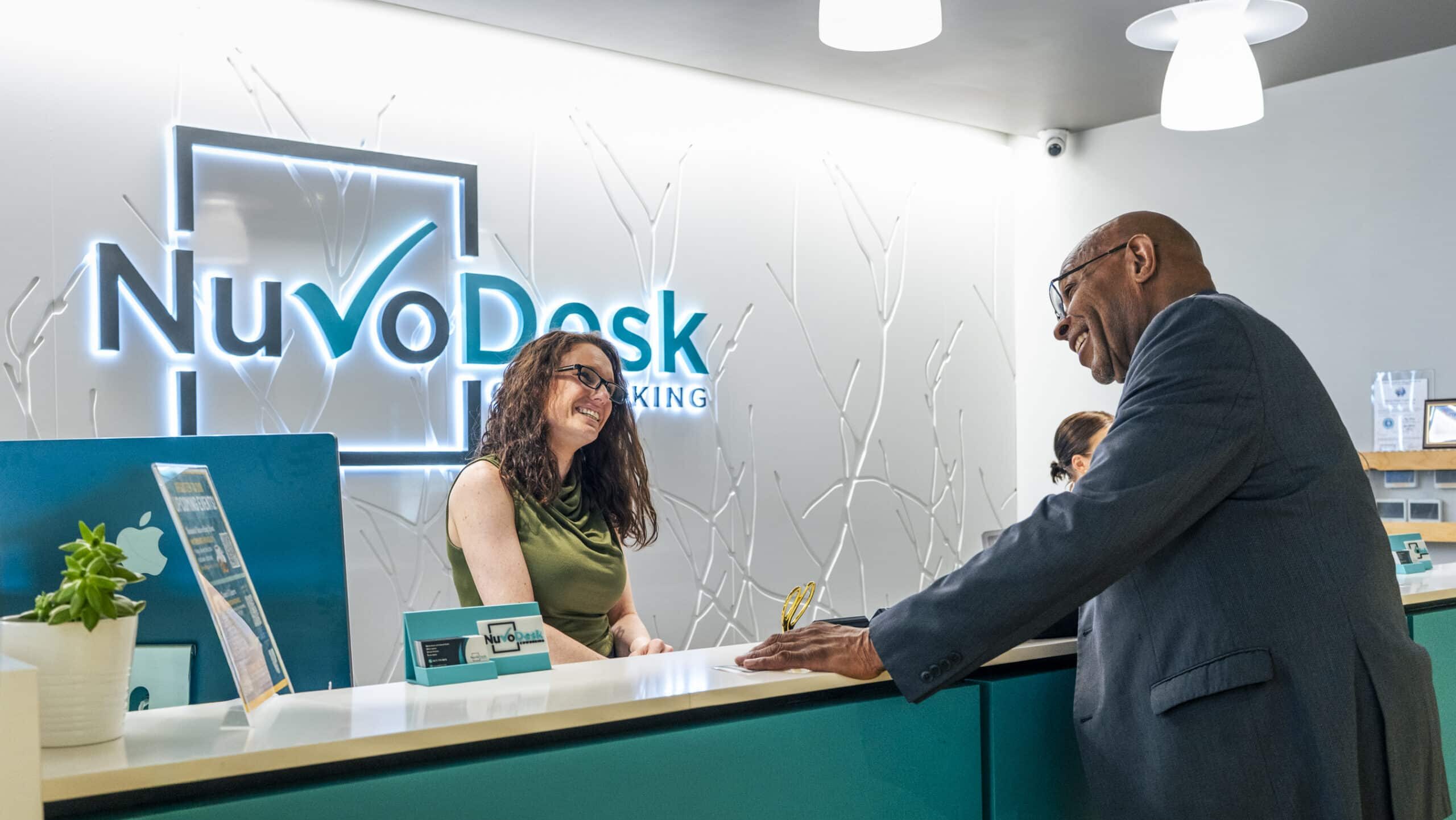 Smiling receptionist in a green top assists a man at a teal front desk with NuVoDesk logo lit on the wall behind them.