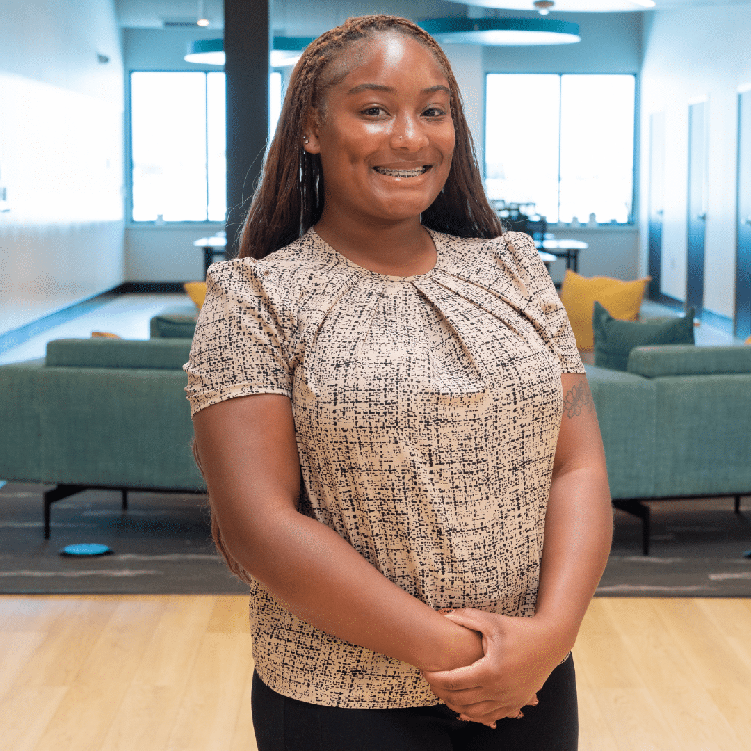 Trissa Terrell ‣ NuvoDesk Coworking ‣ NuvoDesk Coworking Portrait of a smiling woman with braces standing in a bright, modern office lounge with teal couches in the background.